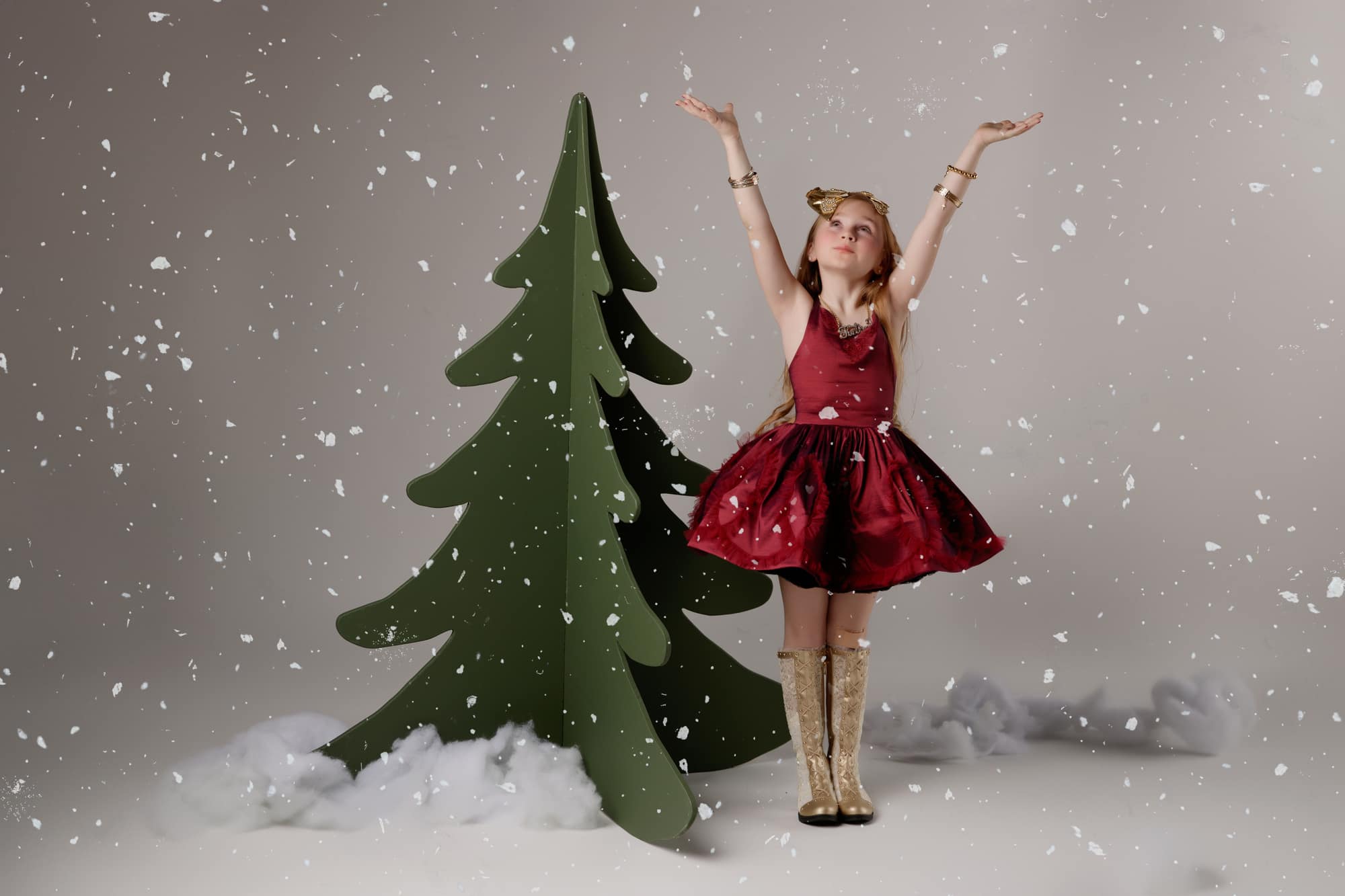 A girl in a red Christmas dress and gold boots reaching up to catch falling snow beside a modern green Christmas tree